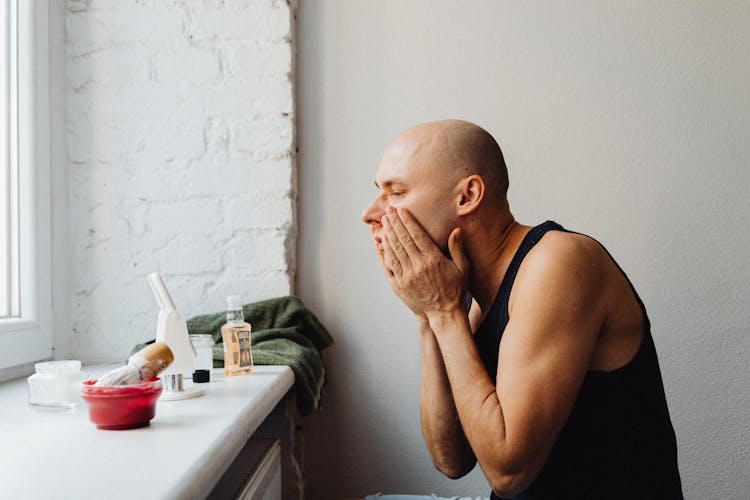 Man Sitting In Front Of A Small Mirror And Using An Aftershave 