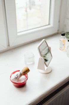 A shaving brush, red container, and mirror on a bright bathroom windowsill.