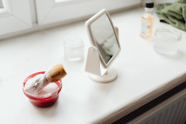 Close-up Of A Mirror And A Container With Shaving Cream And Shaving Brush On A Windowsill 