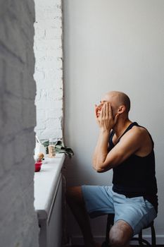 A bald man applies skincare in a relaxed indoor setting by the window.