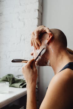 A bald man uses a straight razor and shaving cream in a well-lit room.