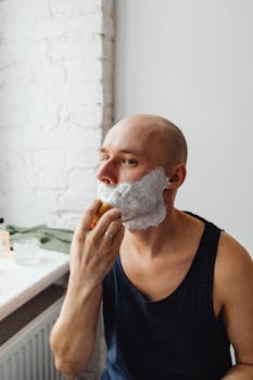 Bald man applying shaving cream with brush in a bathroom setting, demonstrating grooming routine.
