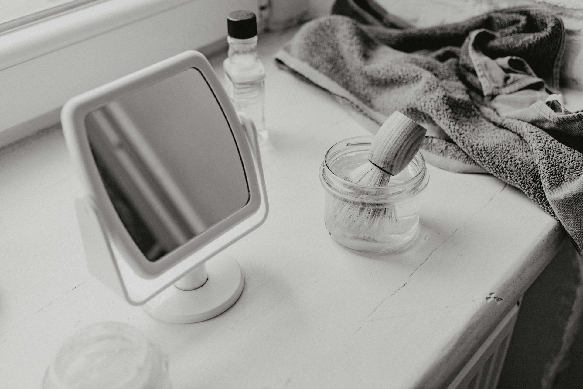 Grayscale Photo of a Mirror and a Brush in a Container