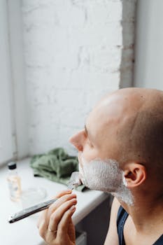 Close-up of a bald man using a straight razor with shaving cream in a bright indoor setting.