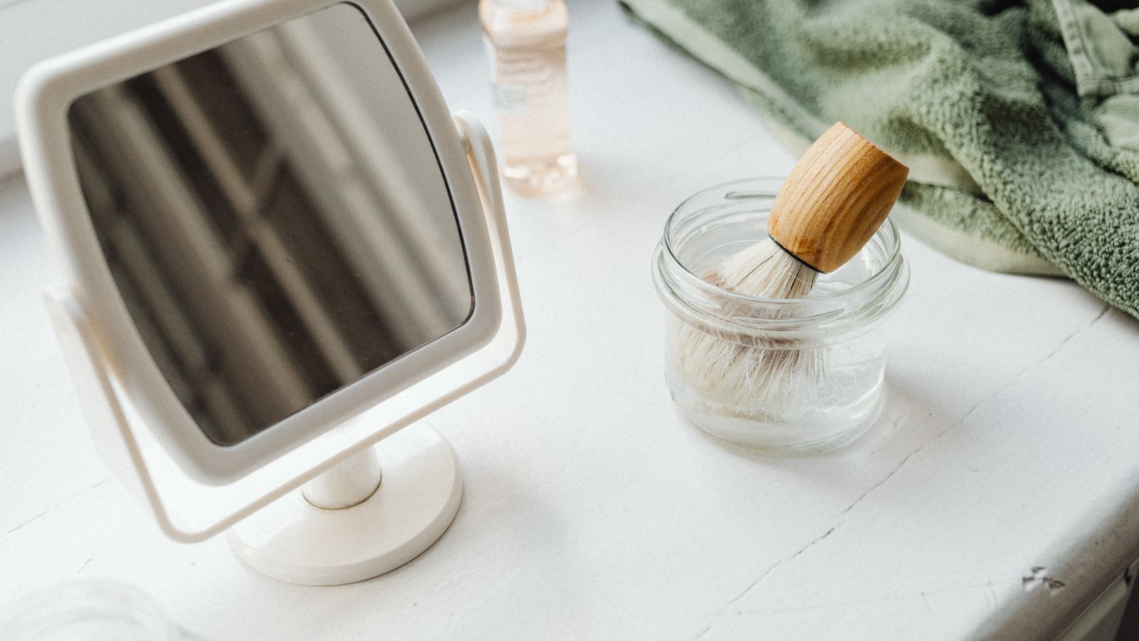 A small mirror and shaving brush on a white surface beside a green towel