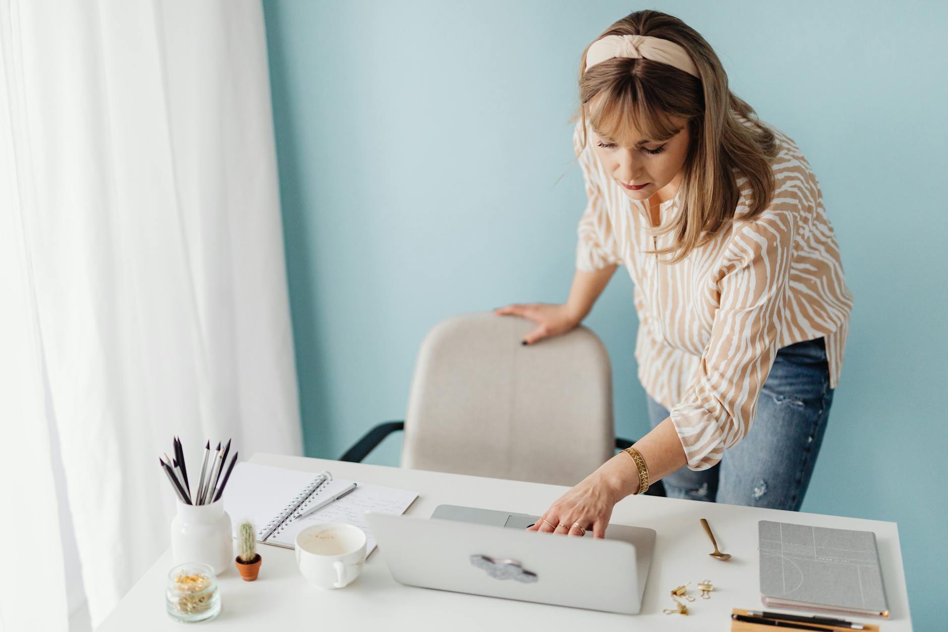 Woman standing at a desk using a laptop with notepad and coffee cup nearby, professional setting.