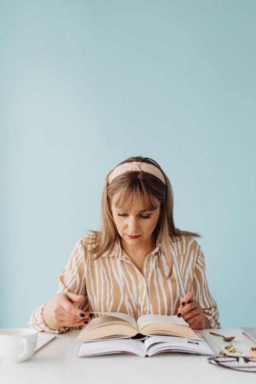 Free Adult woman reading a book at a white table with a light blue background, conveying a peaceful mood. Stock Photo