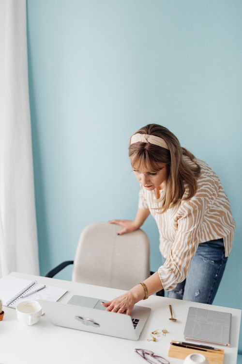 Free Woman in casual attire working on laptop at a minimalist home office desk with a light blue wall. Stock Photo