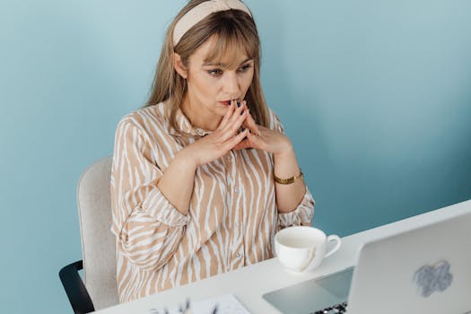 Thoughtful woman at desk with laptop and coffee, in a light blue office setting.