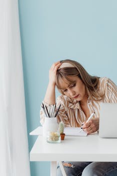 Woman sitting at desk with hand on head, writing in notebook with laptop nearby.