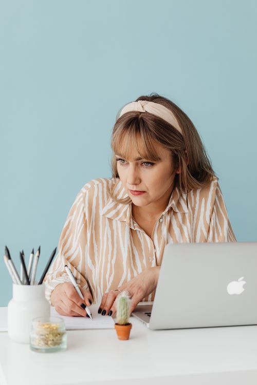 Free Woman in a striped blouse writing at a desk with a laptop and notebook, blue background. Stock Photo
