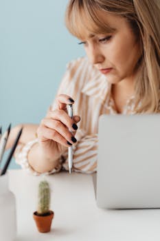 Caucasian woman holding a pen, working at a desk, next to a laptop and small cactus.