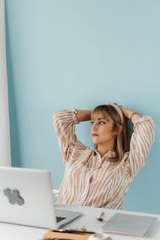 A thoughtful woman leaning back at a desk with a laptop, set against a light blue wall.