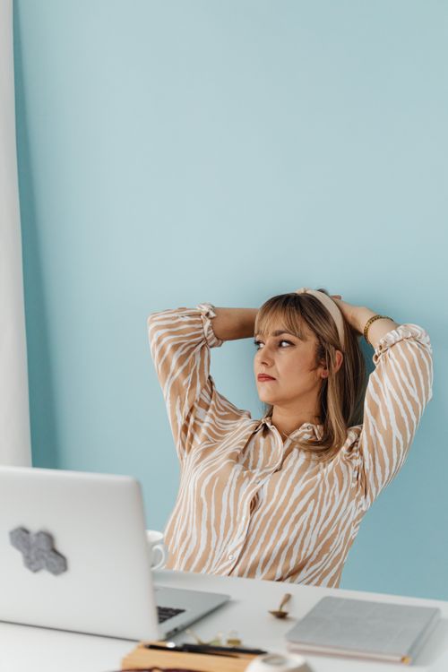 Free A thoughtful woman leaning back at a desk with a laptop, set against a light blue wall. Stock Photo