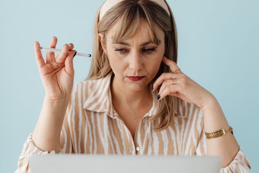 Woman with a pen in hand concentrating on work at her laptop, set against a blue background.