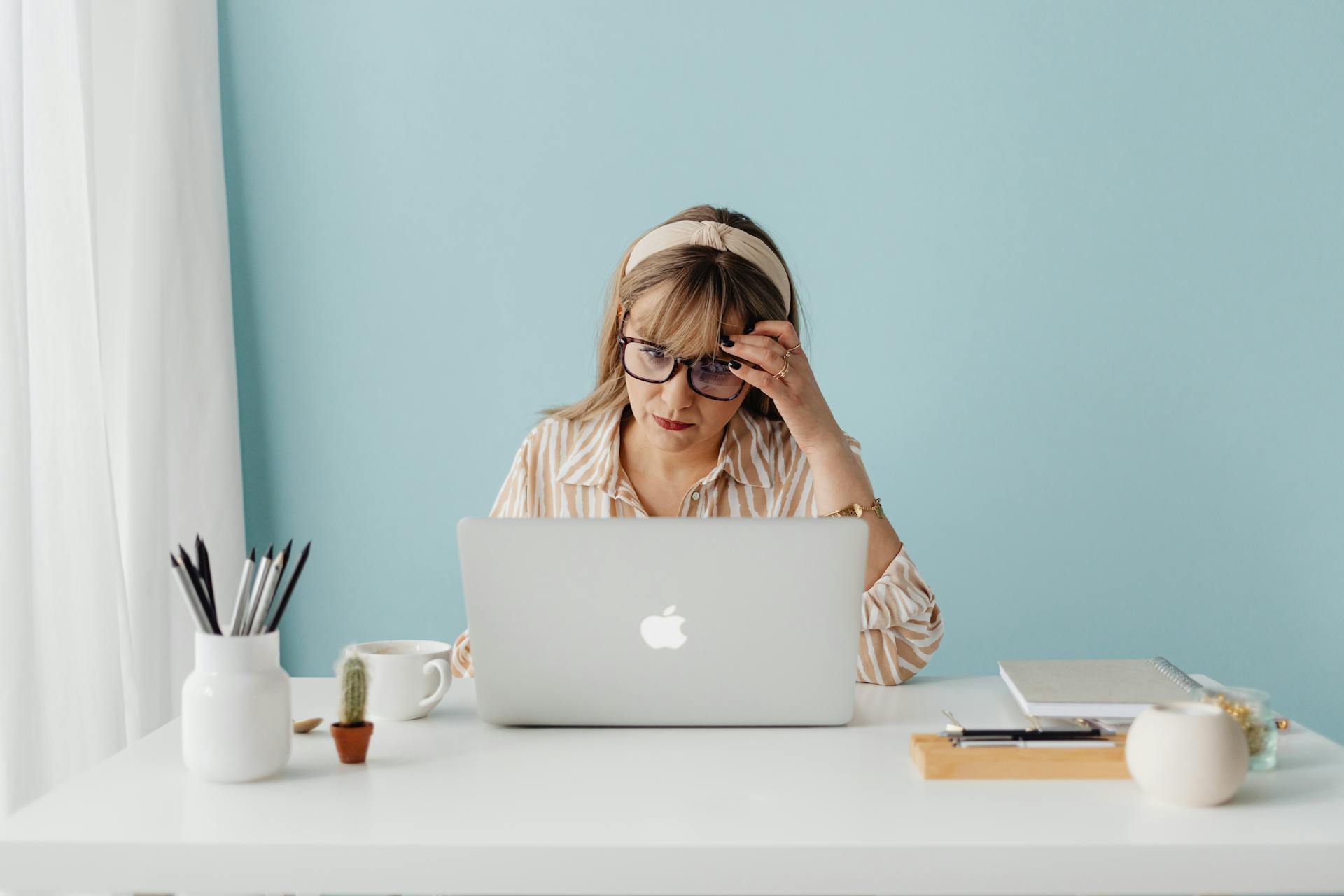 Woman wearing glasses working intently on a laptop at a bright, tidy desk.