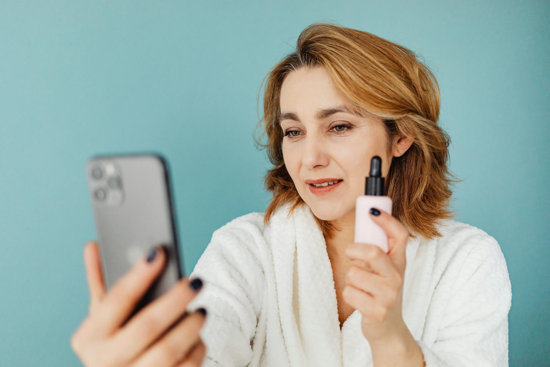 Woman in a white robe taking a selfie while holding a cosmetic bottle against a blue background.