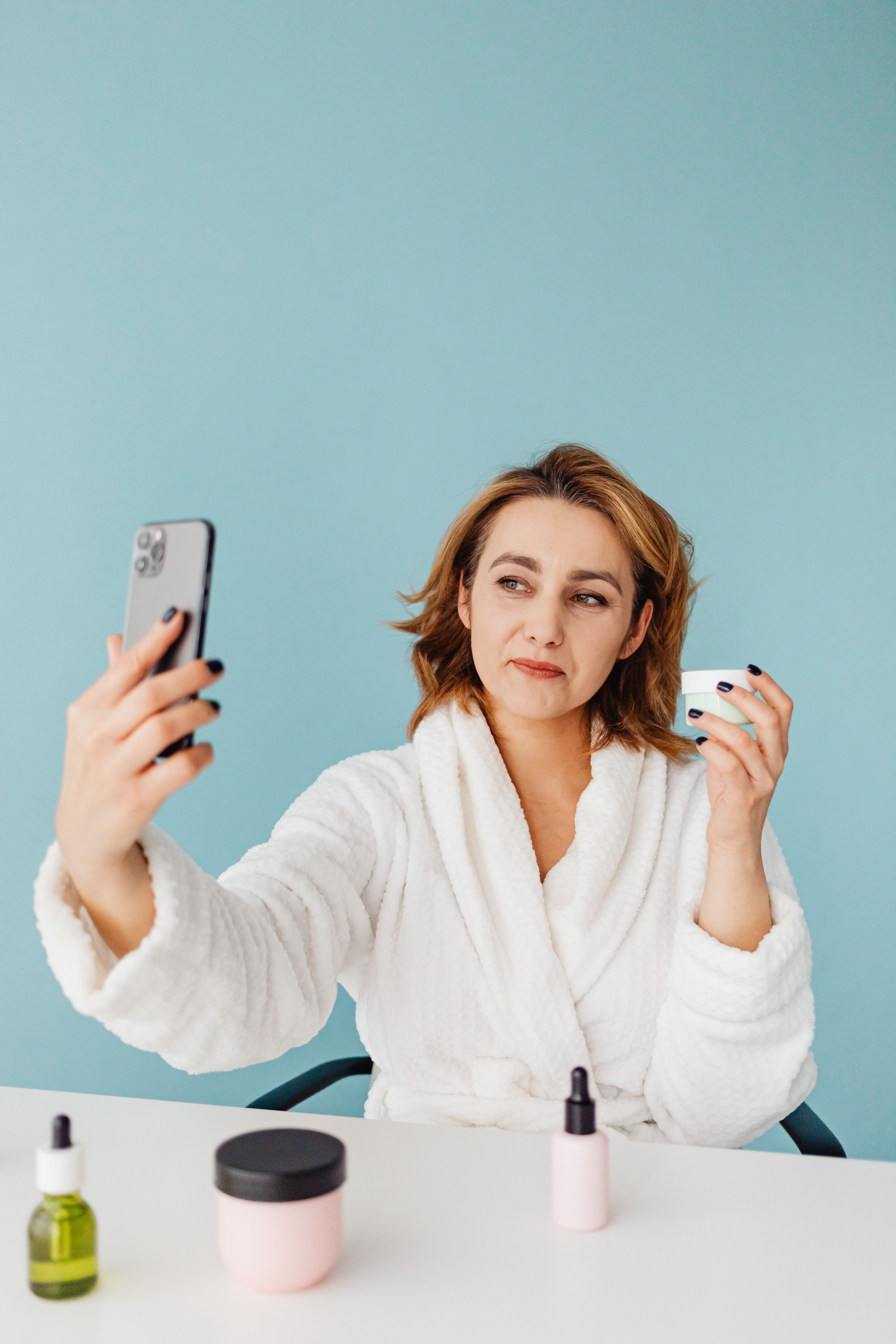 Woman in bathrobe taking selfie holding skincare products on a desk.
