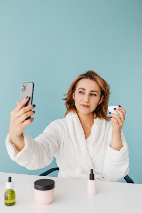 Free Woman in bathrobe taking selfie holding skincare products on a desk. Stock Photo