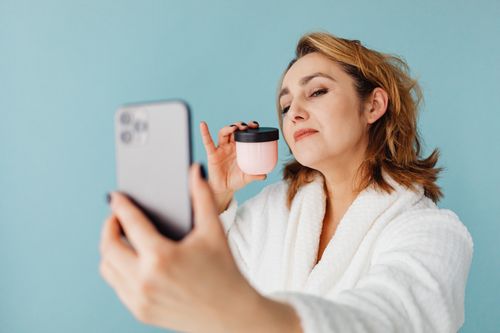 Free Woman in bathrobe taking a selfie holding a skincare product against a blue background. Stock Photo