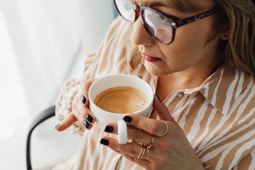 Adult woman wearing eyeglasses savoring a cup of coffee with painted nails indoors.