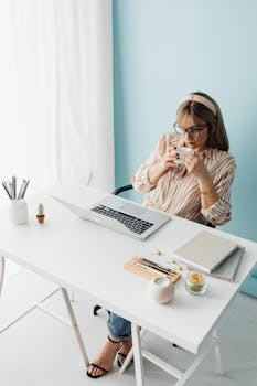 Woman in a striped blouse sitting at a desk with a laptop, enjoying coffee.