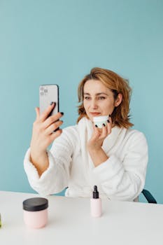 Adult woman in bathrobe taking a smartphone selfie while holding skincare cream indoors.