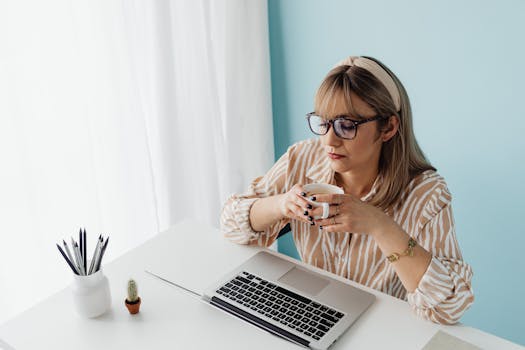 A woman in glasses enjoys a coffee break while working at her desk with a laptop.