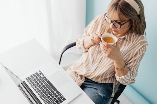 Adult woman sipping coffee and working on laptop in a bright home office setting.
