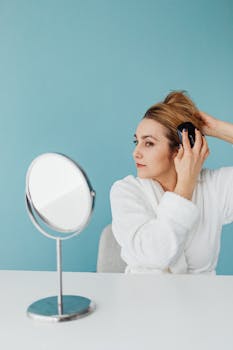 A woman in a bathrobe styles her hair in front of a stand mirror against a blue background.