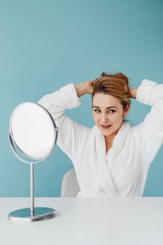 Cheerful woman smiling in a white robe while adjusting her hair in front of a mirror against a light blue background.