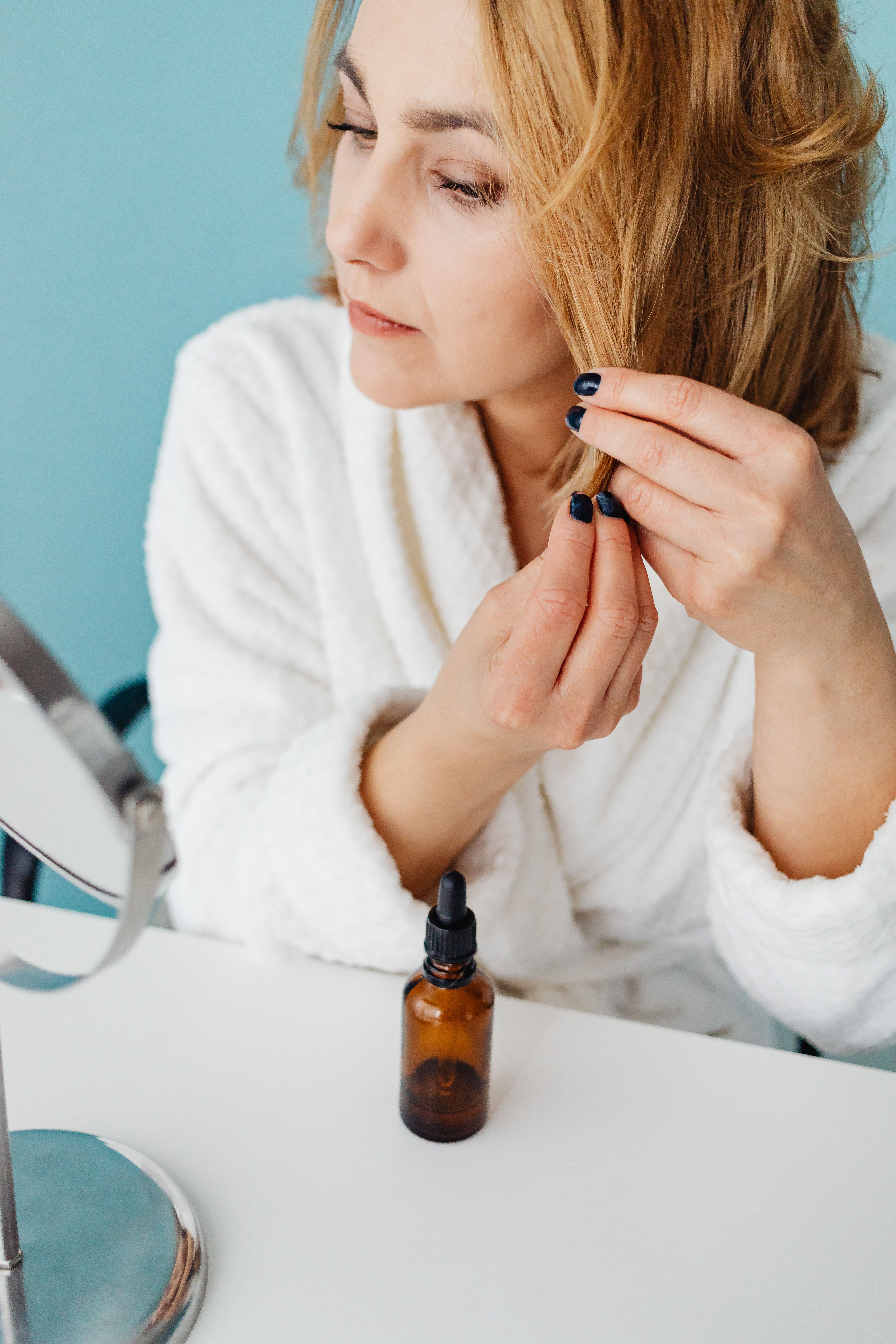 Woman in bathrobe applying hair oil while seated at a table with a mirror.