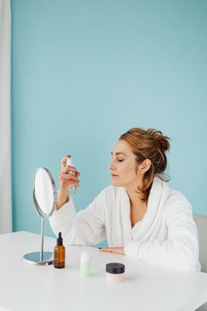 Woman in white bathrobe applying skincare in front of a mirror with closed eyes.
