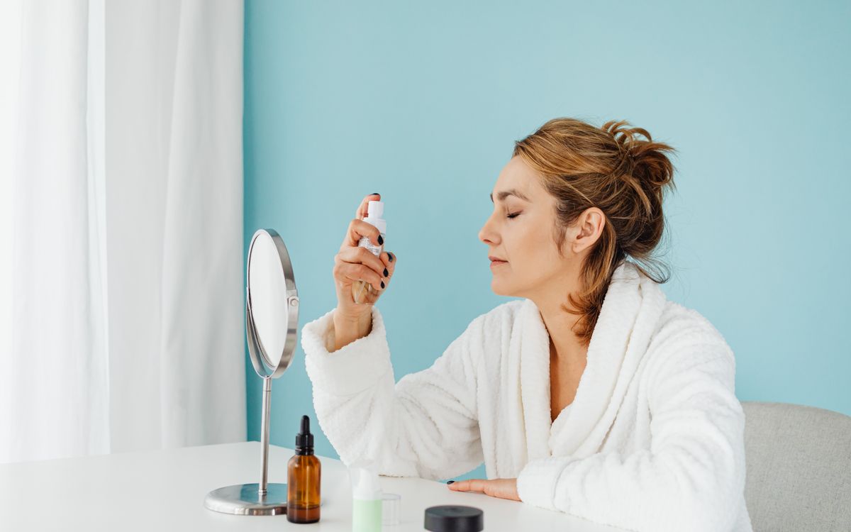 Woman in a bathrobe using a skincare spray in front of a mirror indoors.