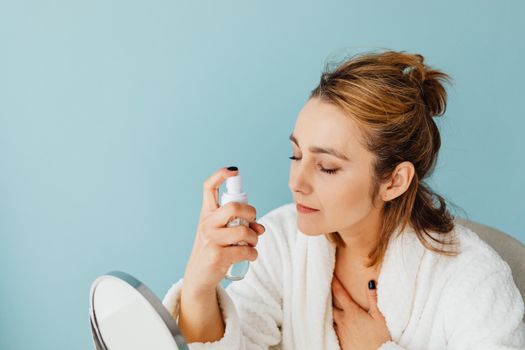 Woman in a bathrobe sprays skincare product onto her face with a blue background.