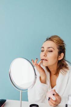 Caucasian woman in white robe applying skincare cream while looking in a mirror.