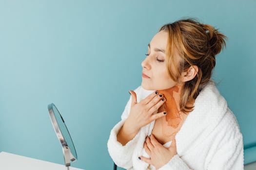 Woman in a white robe applies skincare product in a studio with a blue background.