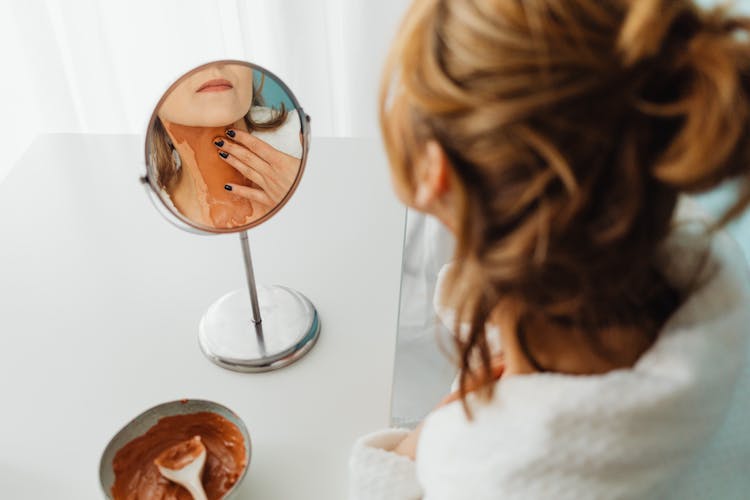 Woman Applying Mud Mask On Her Neck And Reflecting In A Round Mirror