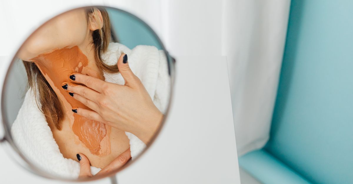 Close-up of a woman applying a thick layer of foundation on her neck in a mirror reflection.