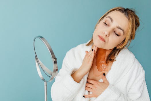 Woman in bathrobe applies skincare treatment using a mirror against a blue backdrop.