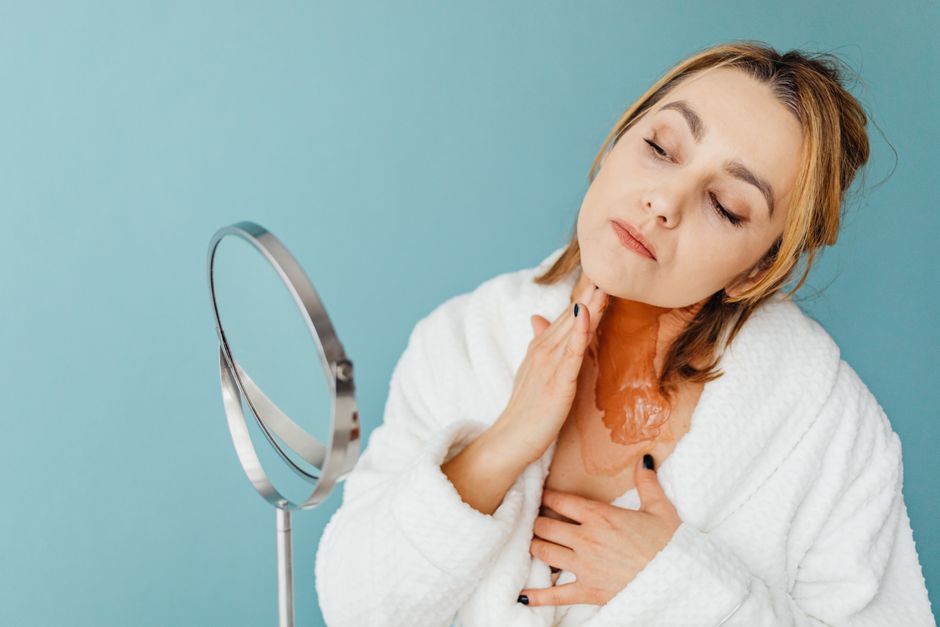 Woman in bathrobe applies skincare treatment using a mirror against a blue backdrop.