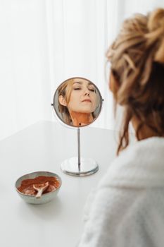 Caucasian woman in robe applies clay mask, reflected in round mirror on white table.