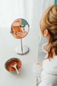 Woman in a bathrobe applying a clay mask to her neck, focused on self-care and beauty routines.