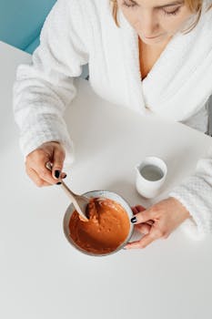Woman in a bathrobe mixing a cosmetic product in a bowl at a table, top-down view.