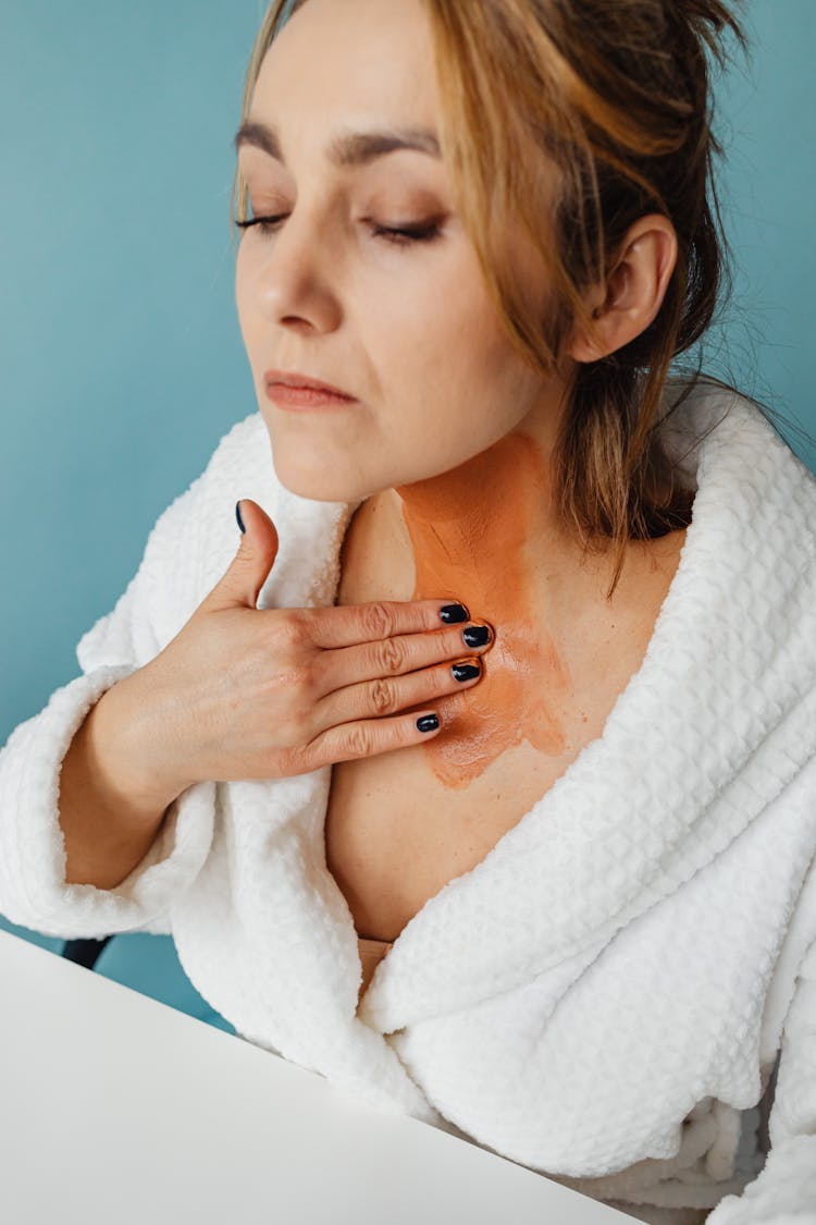 Photo Of A Woman In A White Bathrobe Applying Brown Cream To Her Neck