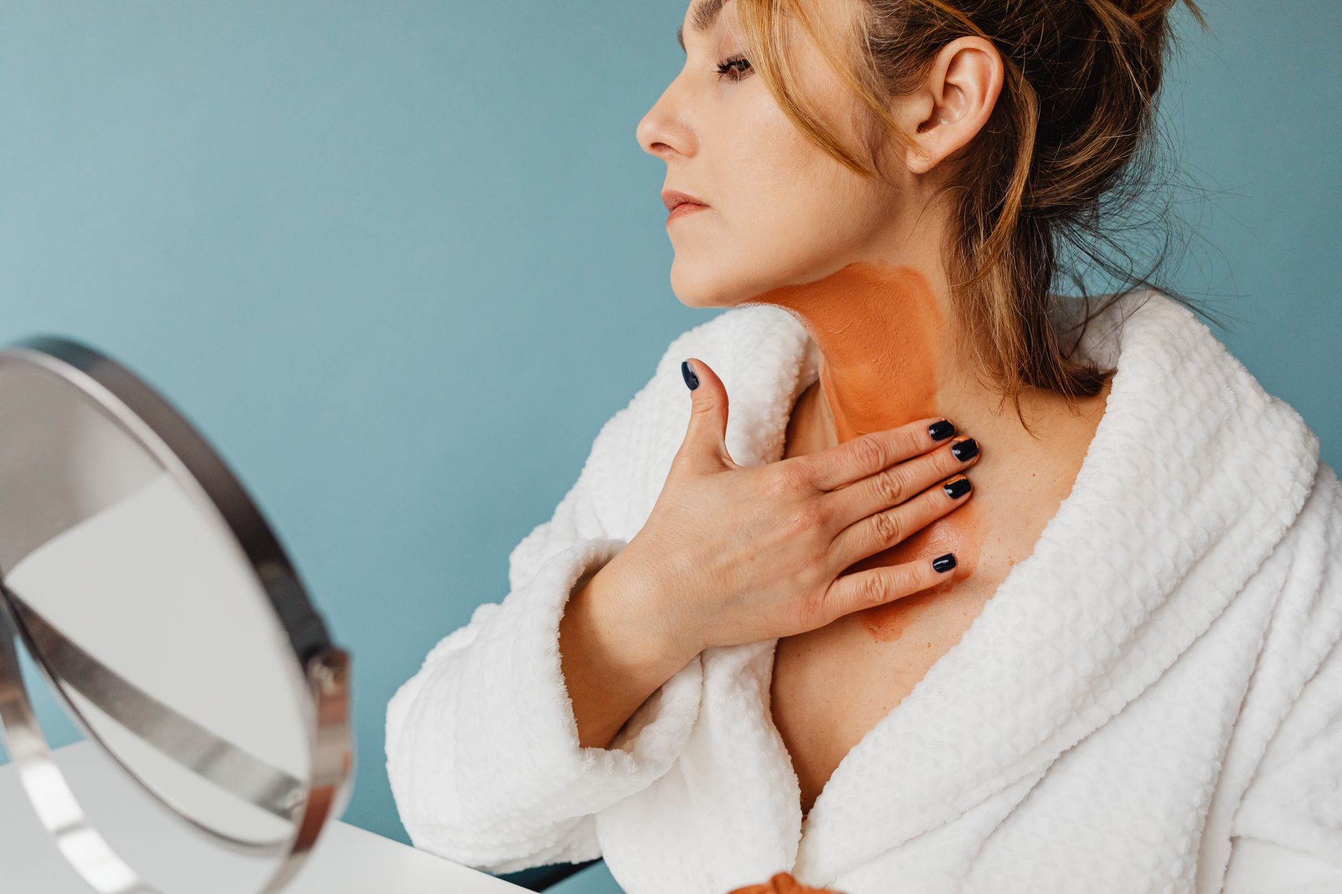 Caucasian woman in bathrobe applying clay mask on neck in indoor setting.
