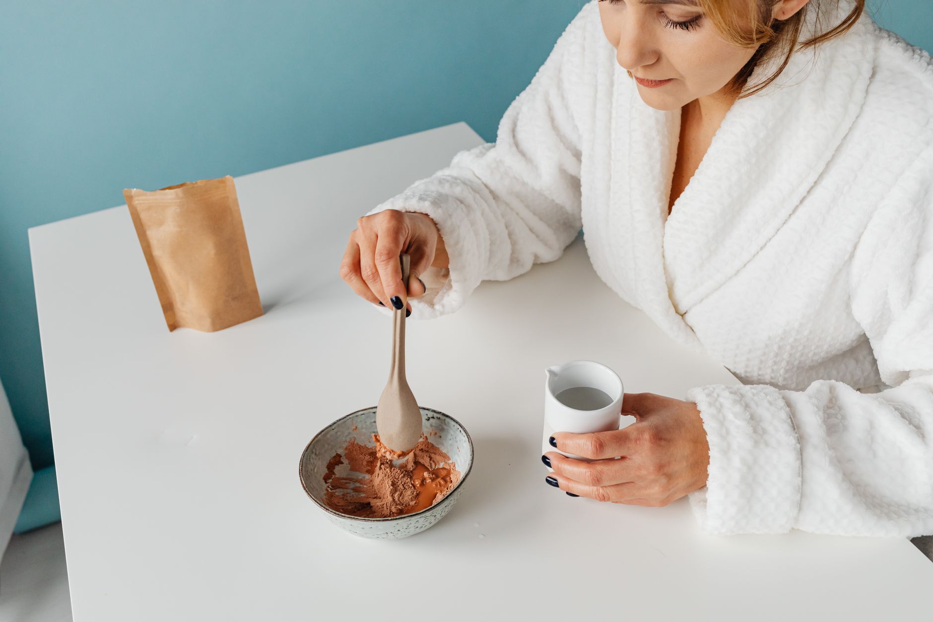 bentonite clay for gut repair after long covid — A woman in a bathrobe mixing a clay face mask with water for skincare purposes.
