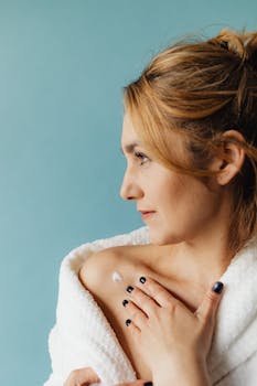 Blonde woman in white robe applying cream to shoulder against blue background.