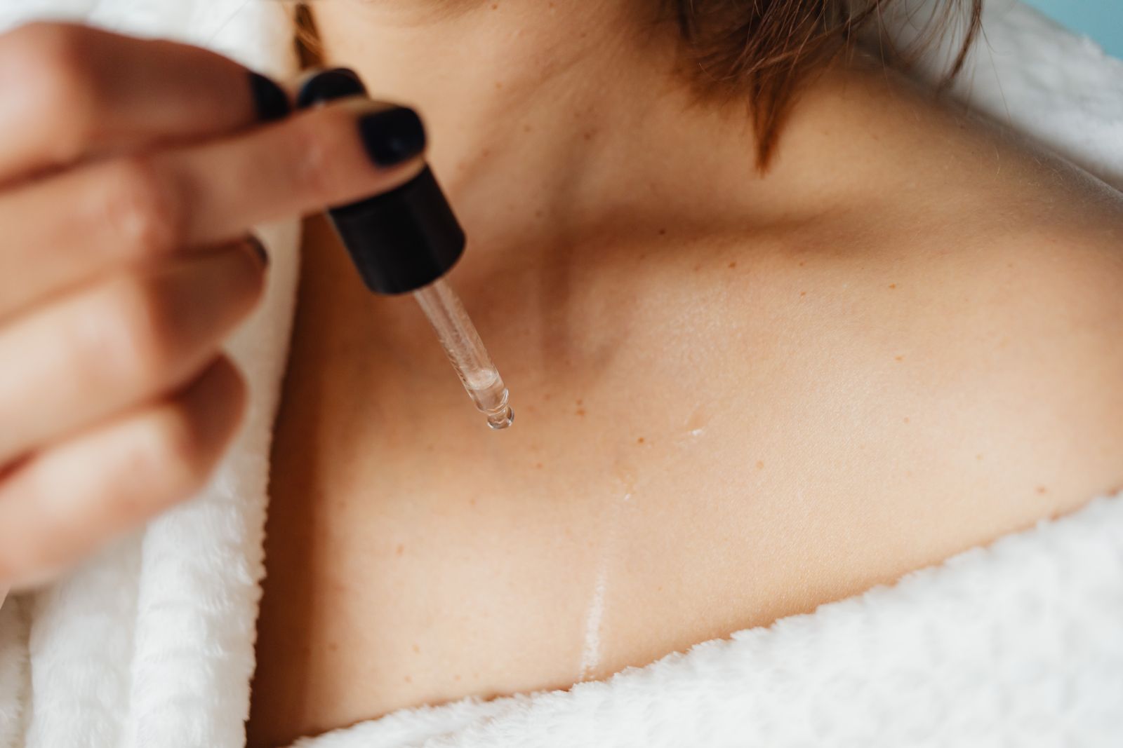 Woman in a white robe applying clear oil from a glass dropper to her collarbone