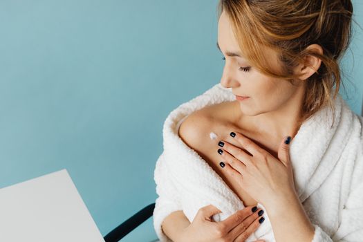Adult woman applying skin cream on shoulder while wearing a bathrobe against a blue background.